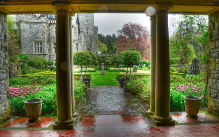 A picturesque view from beneath columns into a lush courtyard garden, featuring blooming flowers and a charming castle in the background, creating a serene, man-made landscape.