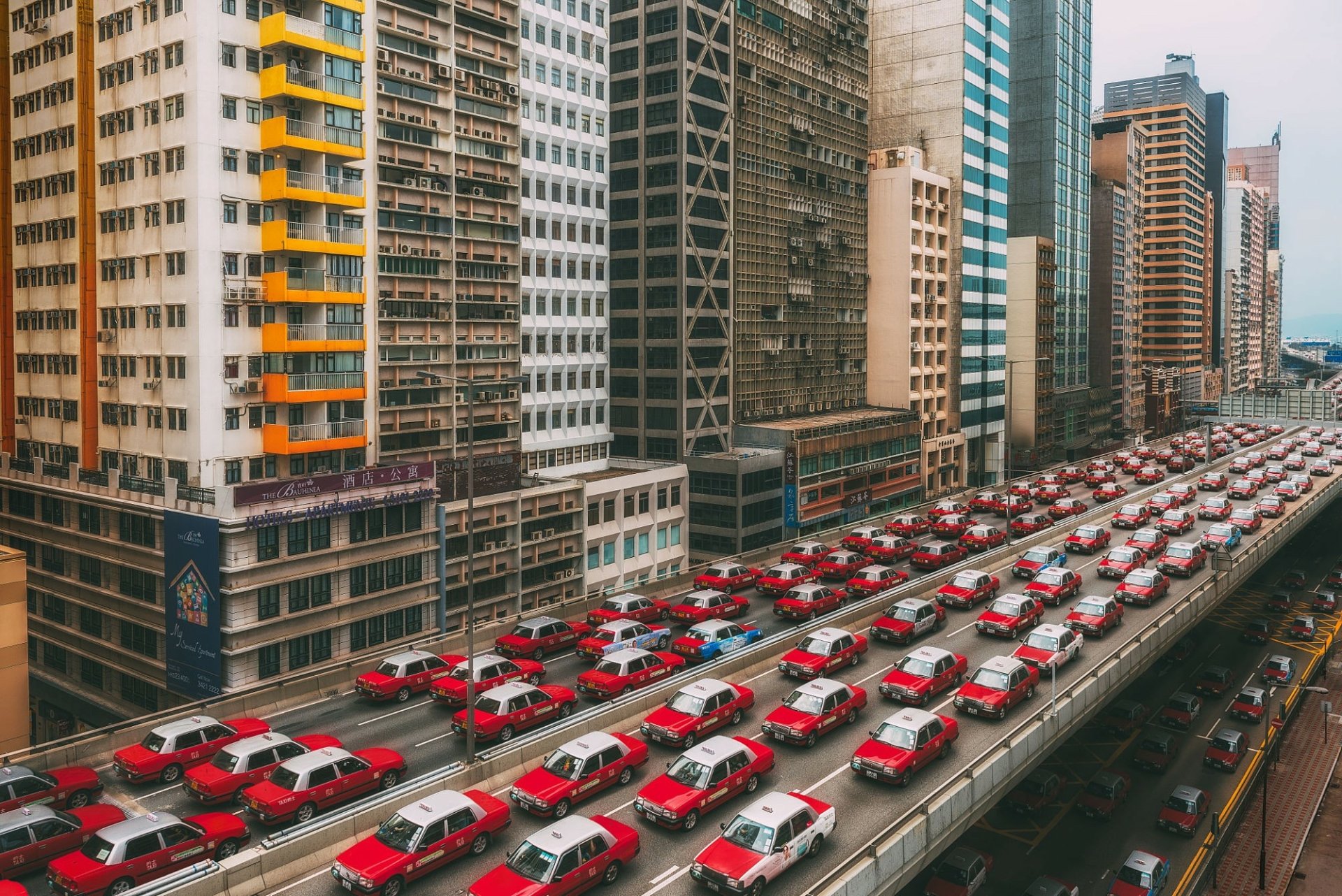 A bustling Hong Kong street filled with iconic red taxis lined up along a busy highway flanked by tall, densely packed buildings in this HD desktop wallpaper.