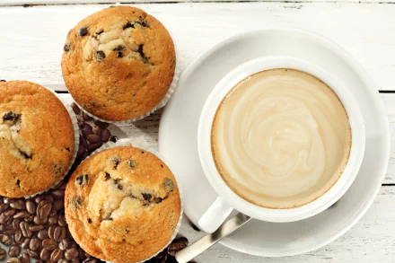 A 4K Ultra HD desktop wallpaper featuring three chocolate chip muffins beside a white cup of creamy coffee on a saucer, set on a white wooden surface with coffee beans.