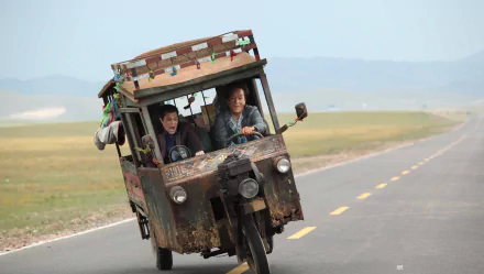 Jackie Chan and a young boy joyfully ride in a rustic vehicle on an open road in a scene from the movie Skiptrace (2016). A vibrant HD desktop wallpaper for fans.