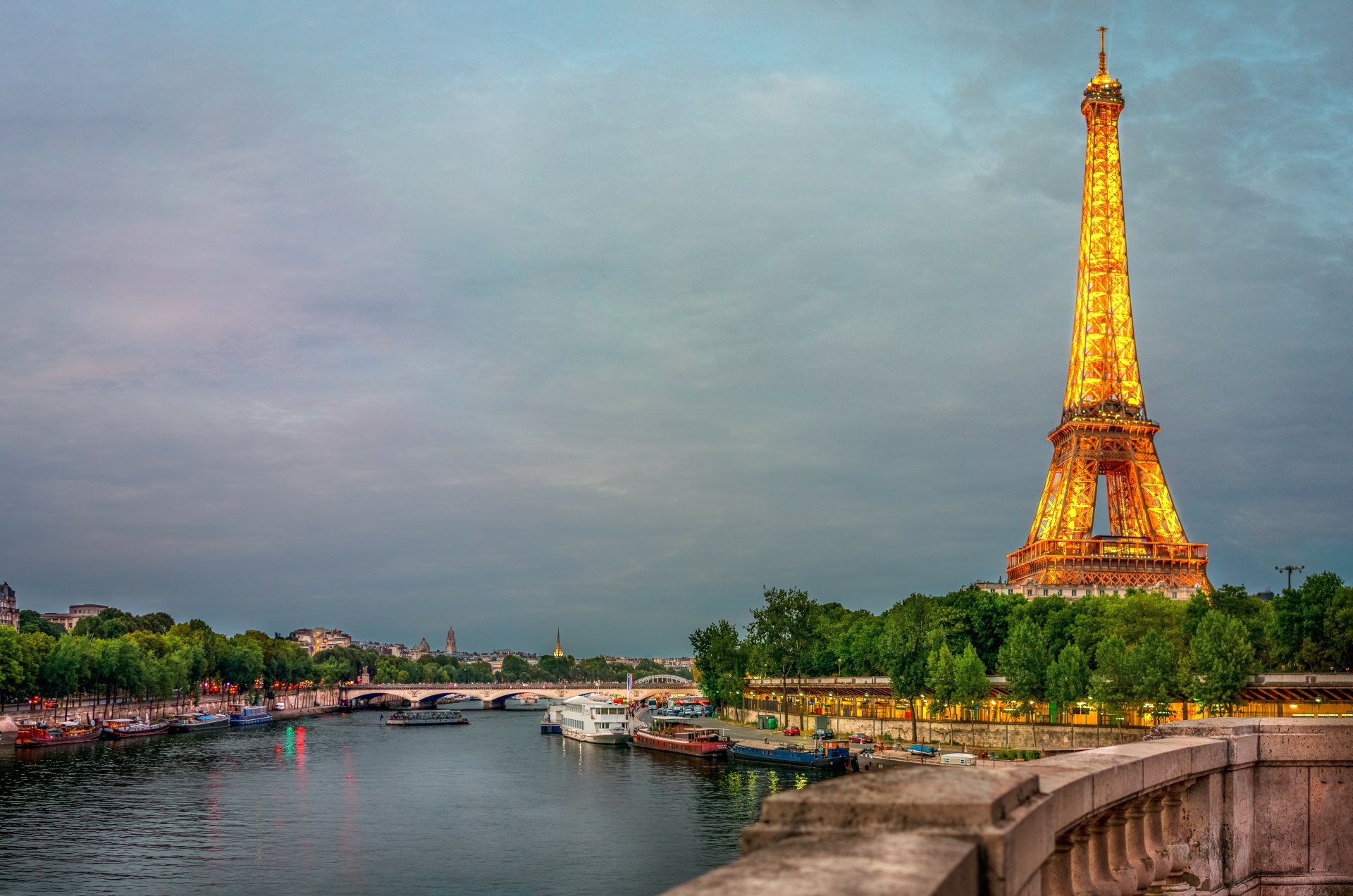 8K Ultra HD view of the Eiffel Tower glowing at dusk beside the Seine River in Paris, with boats docked along the water and a stone bridge in the foreground.
