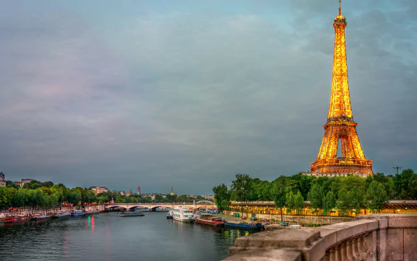 8K Ultra HD view of the Eiffel Tower glowing at dusk beside the Seine River in Paris, with boats docked along the water and a stone bridge in the foreground.
