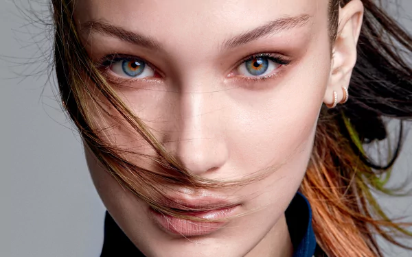 Close-up HD desktop wallpaper of a brunette female model with striking blue eyes, wind-swept hair across her face and a subtle gold hoop earring.