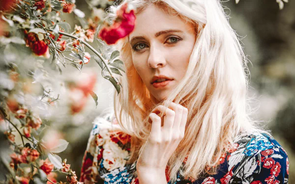5K Ultra HD desktop wallpaper of a blonde, blue‑eyed woman model outdoors with soft bokeh and roses in a floral background, contemplative expression.