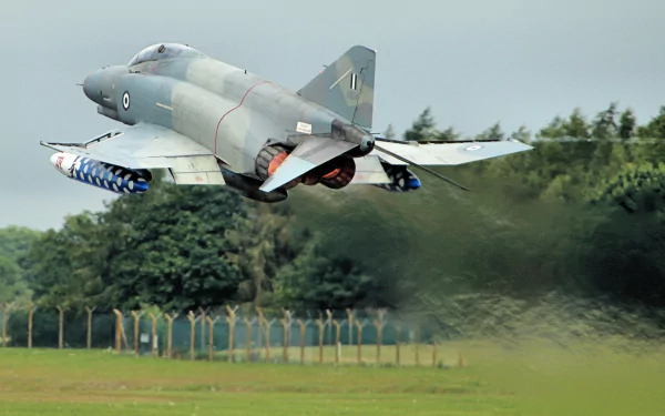 McDonnell Douglas F-4 Phantom II jet fighter warplane taking off over a grassy field with trees in the background, captured in high definition.