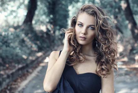 HD wallpaper featuring a brunette woman with curly hair, set against a bokeh background of a forest path. The model has a serene expression and is dressed in a strapless black top.
