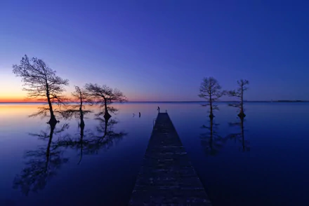 HD desktop wallpaper showing a tranquil lake at sunset with a wooden pier extending toward the horizon, silhouetted trees, and clear reflections under a deep blue sky.