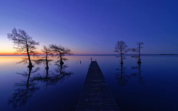 HD desktop wallpaper showing a tranquil lake at sunset with a wooden pier extending toward the horizon, silhouetted trees, and clear reflections under a deep blue sky.