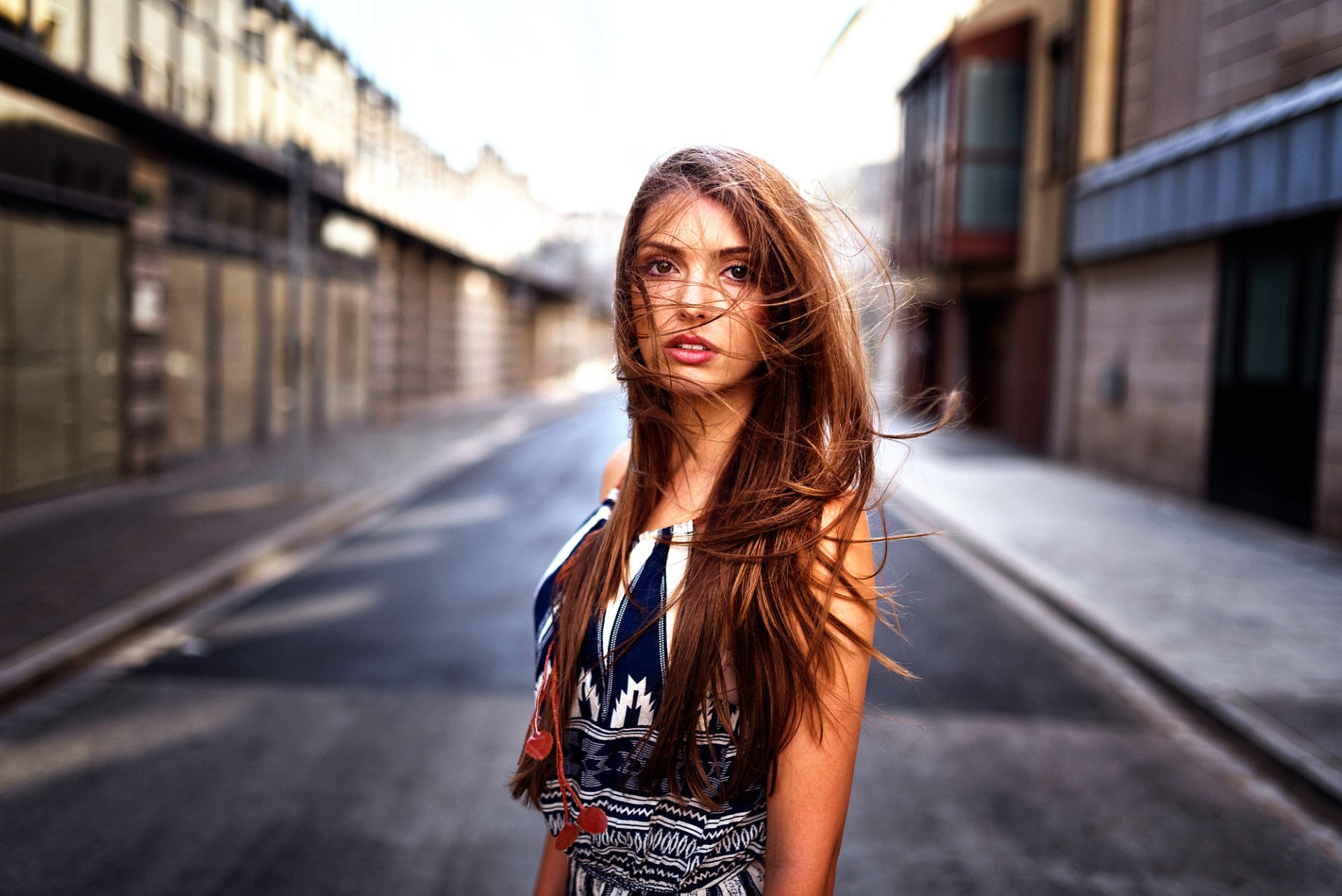 HD desktop wallpaper featuring a brunette woman with brown eyes standing in the middle of a sunlit urban street, with her hair blowing gently in the breeze.