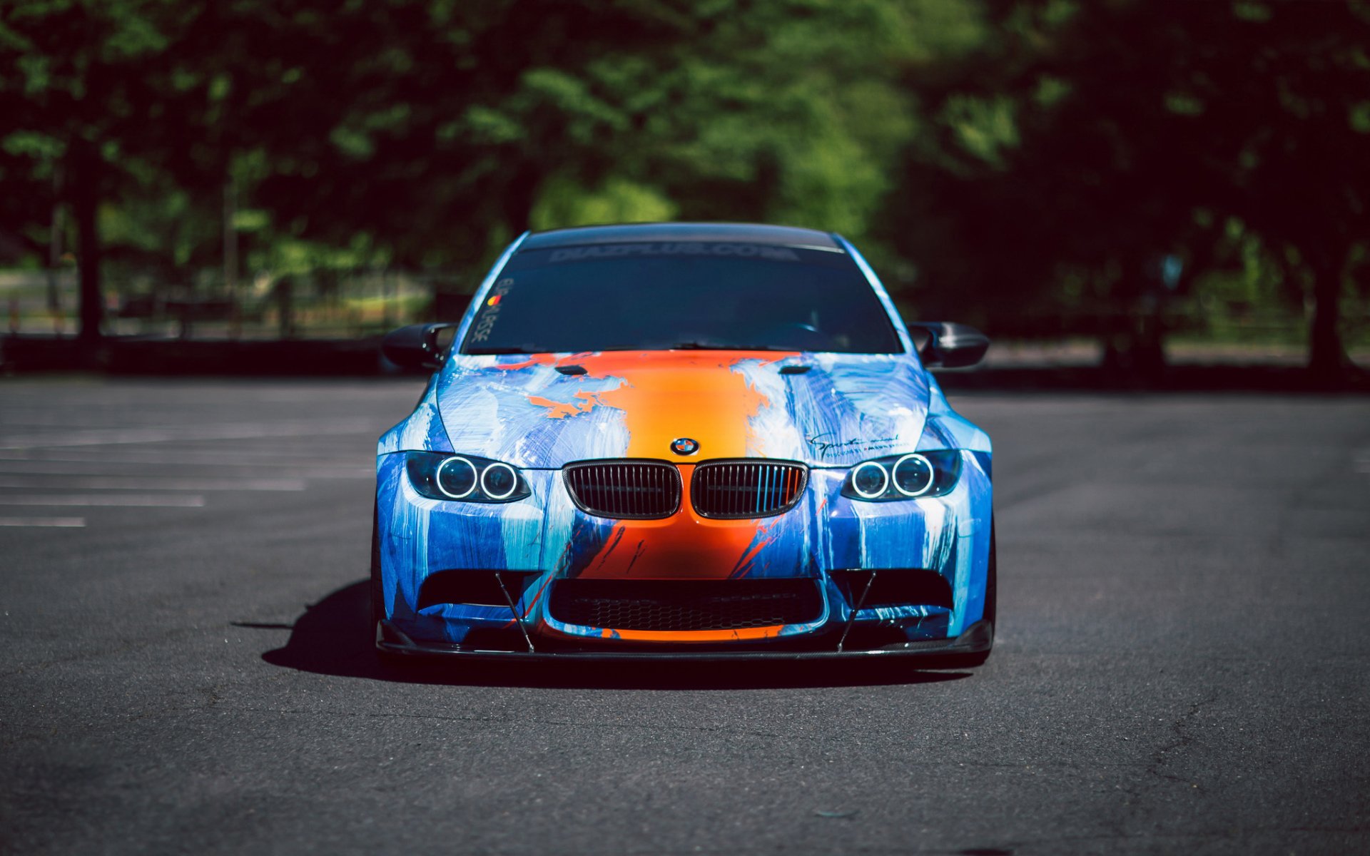 HD desktop wallpaper featuring a BMW M3 with a striking blue and orange paint job, parked on a dark asphalt surface, against a backdrop of green trees.