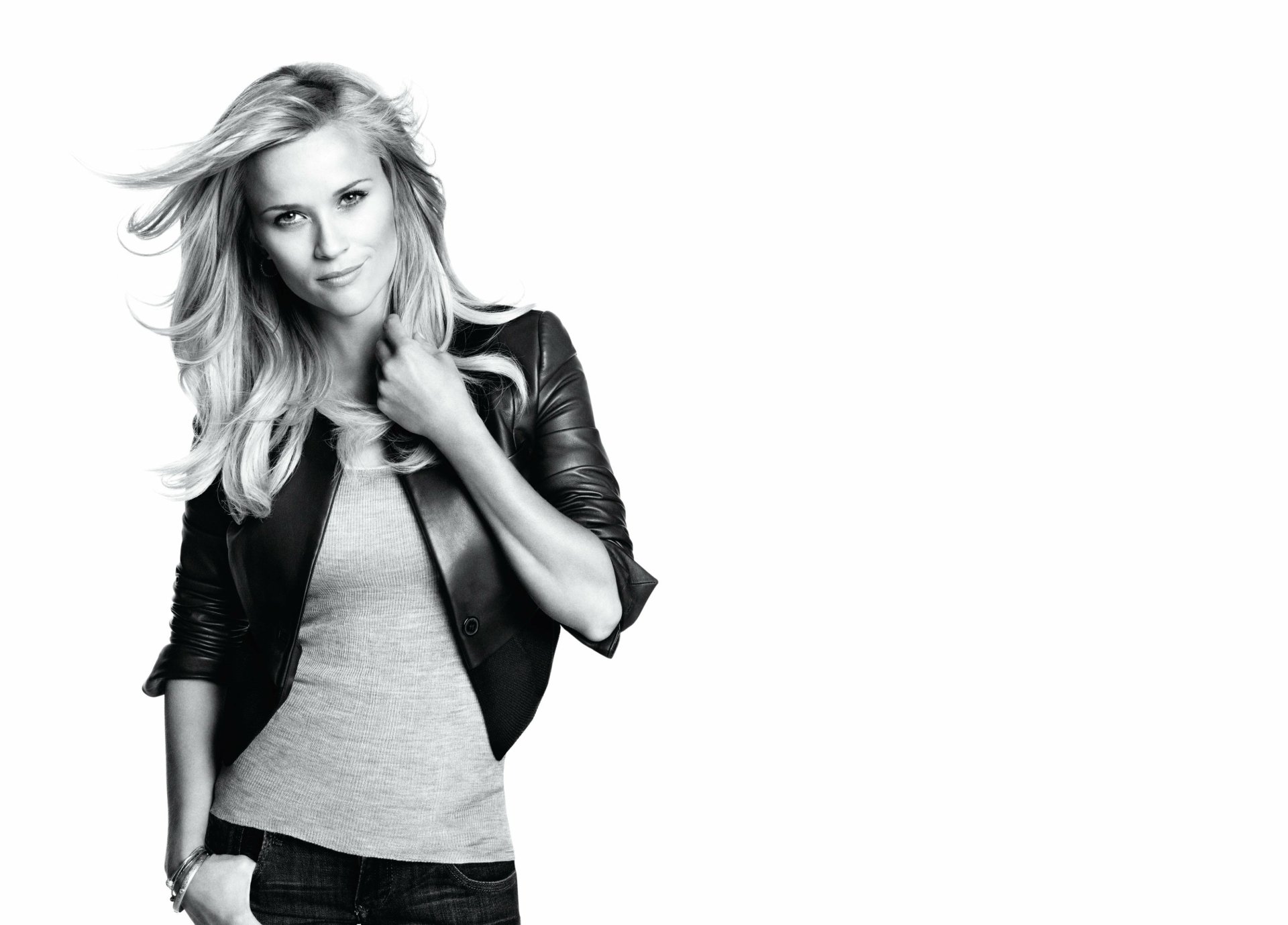 Black-and-white 2K desktop wallpaper: half-length portrait of a blonde woman in a leather jacket, hand at her collar, looking toward the camera against a white background.