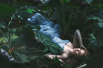 A woman in a flowing blue dress lies among large green leaves, creating a serene and moody atmosphere in this HD desktop wallpaper.