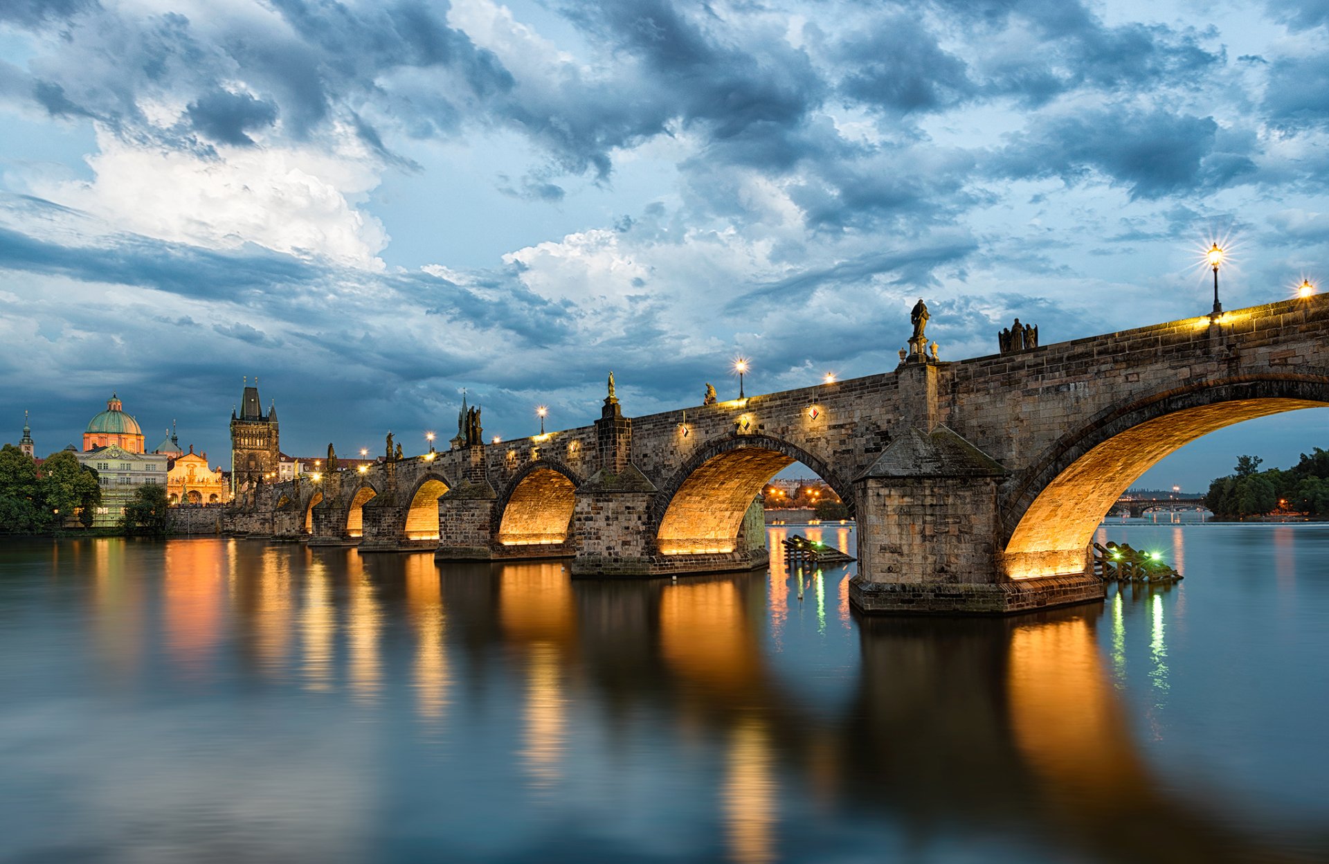 HD desktop wallpaper of Charles Bridge in Prague, Czech Republic at dusk: illuminated man-made stone bridge arches reflected in the calm river with dramatic clouds and glowing lights.