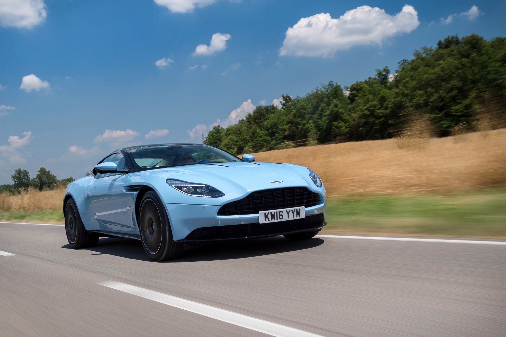A stunning light blue Aston Martin DB11 grand tourer speeds along a scenic road under a bright blue sky in this 4K Ultra HD desktop wallpaper.