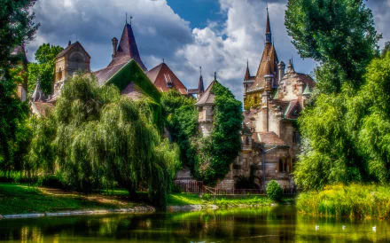 HDR image of a man-made castle in Budapest, Hungary, reflected in a tranquil pond surrounded by lush greenery under a partly cloudy sky.