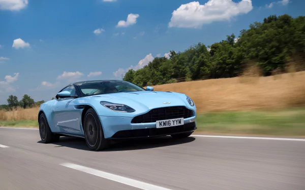 A stunning light blue Aston Martin DB11 grand tourer speeds along a scenic road under a bright blue sky in this 4K Ultra HD desktop wallpaper.