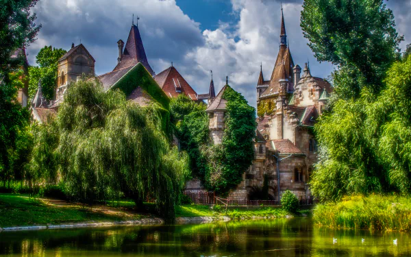 HDR image of a man-made castle in Budapest, Hungary, reflected in a tranquil pond surrounded by lush greenery under a partly cloudy sky.