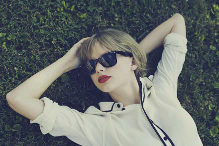 A stylish American singer with blonde hair, wearing sunglasses and bold lipstick, relaxes on the grass, creating a vibrant HD desktop wallpaper.