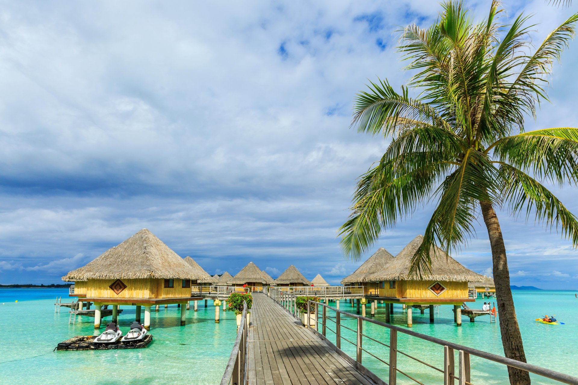 Tropical bungalow over turquoise water with palm trees under a partly cloudy sky in Bora Bora, French Polynesia, captured in a vibrant 4K Ultra HD holiday photograph.