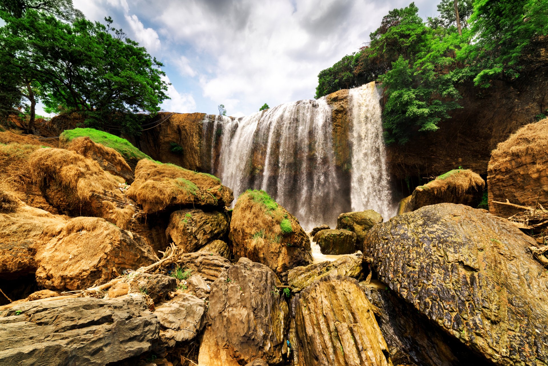 A 4K Ultra HD desktop wallpaper of a vibrant waterfall surrounded by lush greenery and rocks in Vietnam under a partly cloudy sky.
