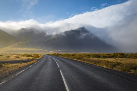 HD desktop wallpaper of a winding man-made road through Iceland’s mountainous landscape, with low clouds partially covering the peaks under a bright sky.