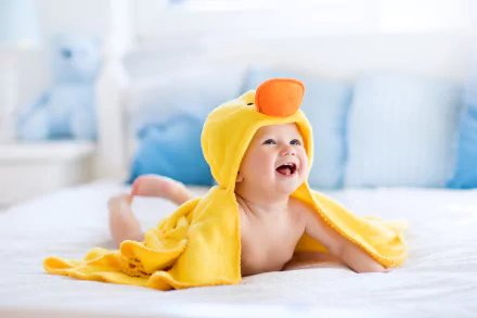 A smiling baby wrapped in a yellow duck towel lies on a bed, surrounded by soft bedding. This image serves as a vivid 8K Ultra HD wallpaper and background.