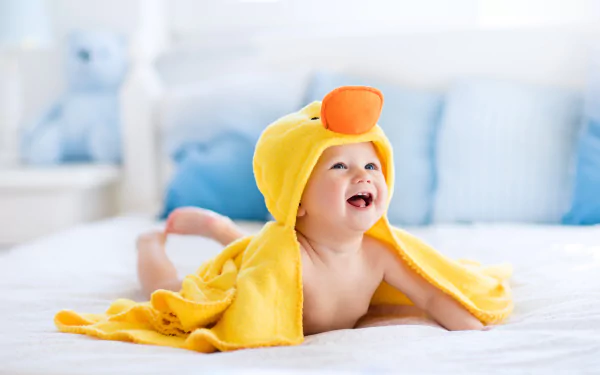 A smiling baby wrapped in a yellow duck towel lies on a bed, surrounded by soft bedding. This image serves as a vivid 8K Ultra HD wallpaper and background.