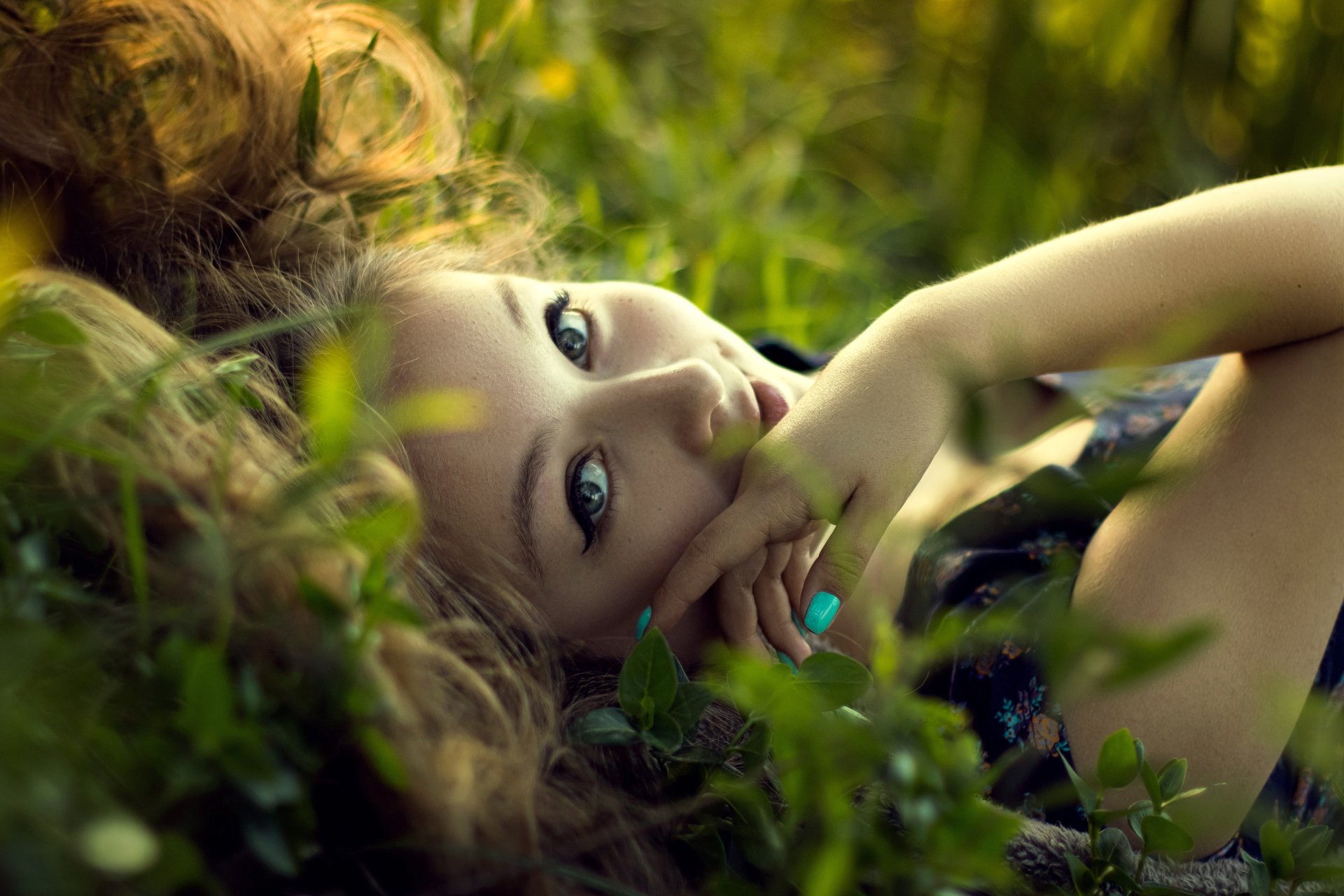 A high-definition wallpaper featuring a woman lying down on grass, gazing up with a serene expression. The close-up emphasizes her face and natural surroundings.