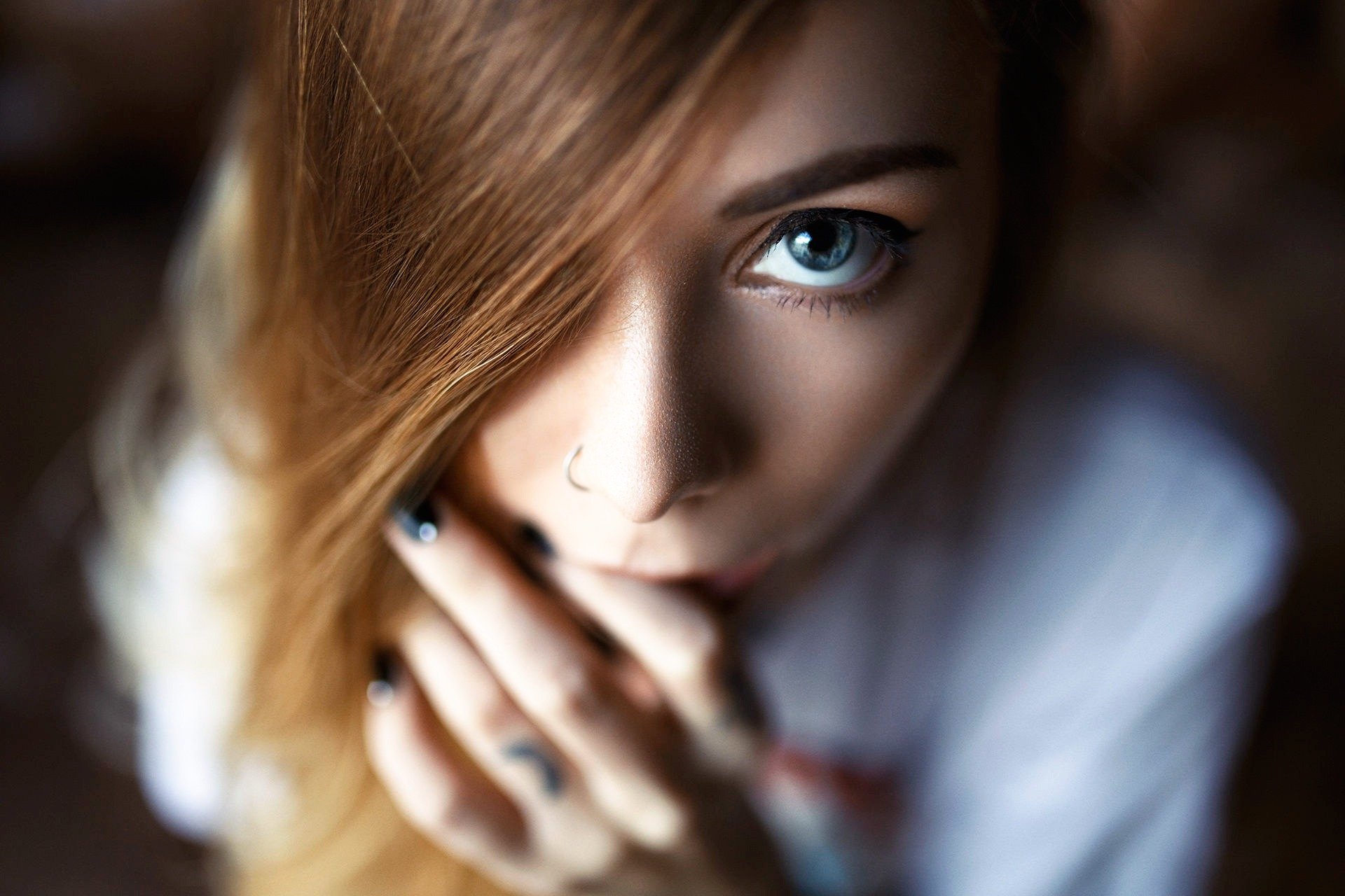 A close-up of a woman with blue eyes and long, flowing hair, her face partially obscured by her hand. The image features a soft blur, creating an intriguing HD wallpaper effect.