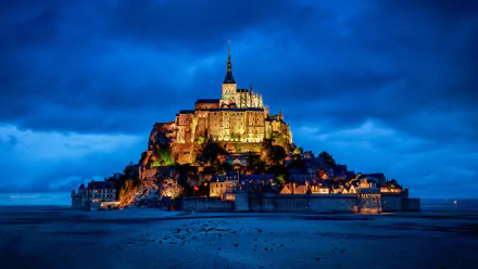Mont Saint-Michel monastery in France illuminated at night, set against a deep blue sky, showcasing its historic religious architecture on a rocky island.