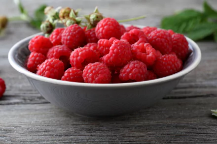 A close-up 4K Ultra HD image of fresh red raspberries in a gray bowl on a wooden surface, highlighting the vibrant color and texture of the fruit and berries.