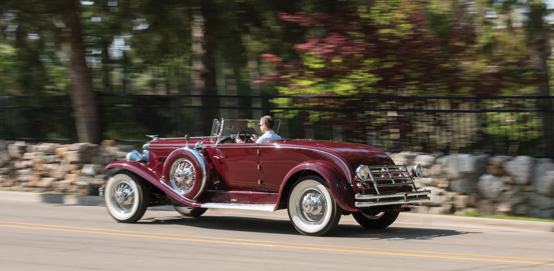A deep red 1929 Duesenberg Model J Disappearing Top Convertible Coupe drives past trees and flowers, showcased as a vintage car in this HD desktop wallpaper.