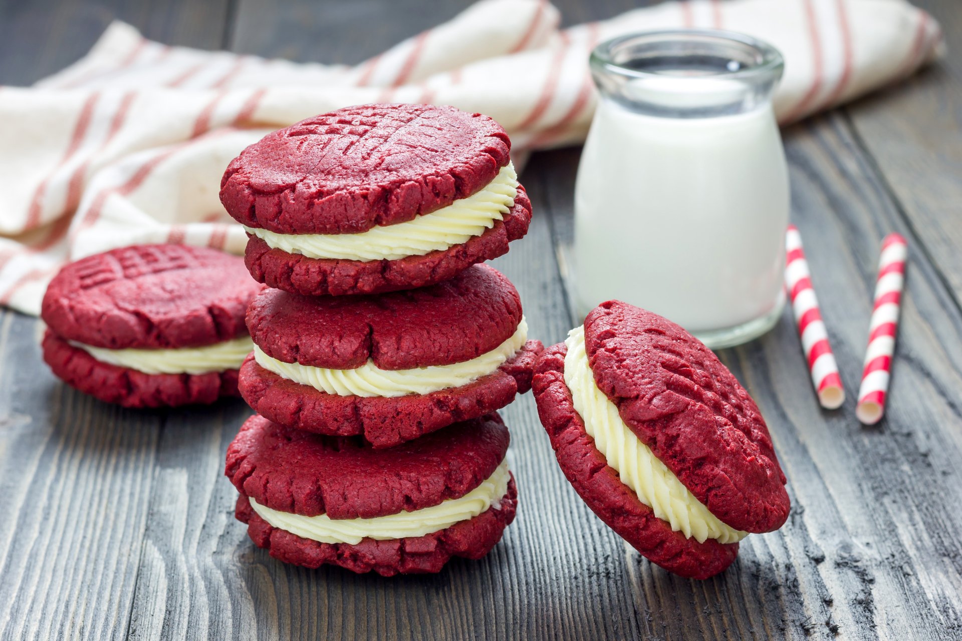 A stack of red velvet biscuits filled with cream stands beside a glass of milk and striped straws, creating a charming food scene in this 4K Ultra HD desktop wallpaper.