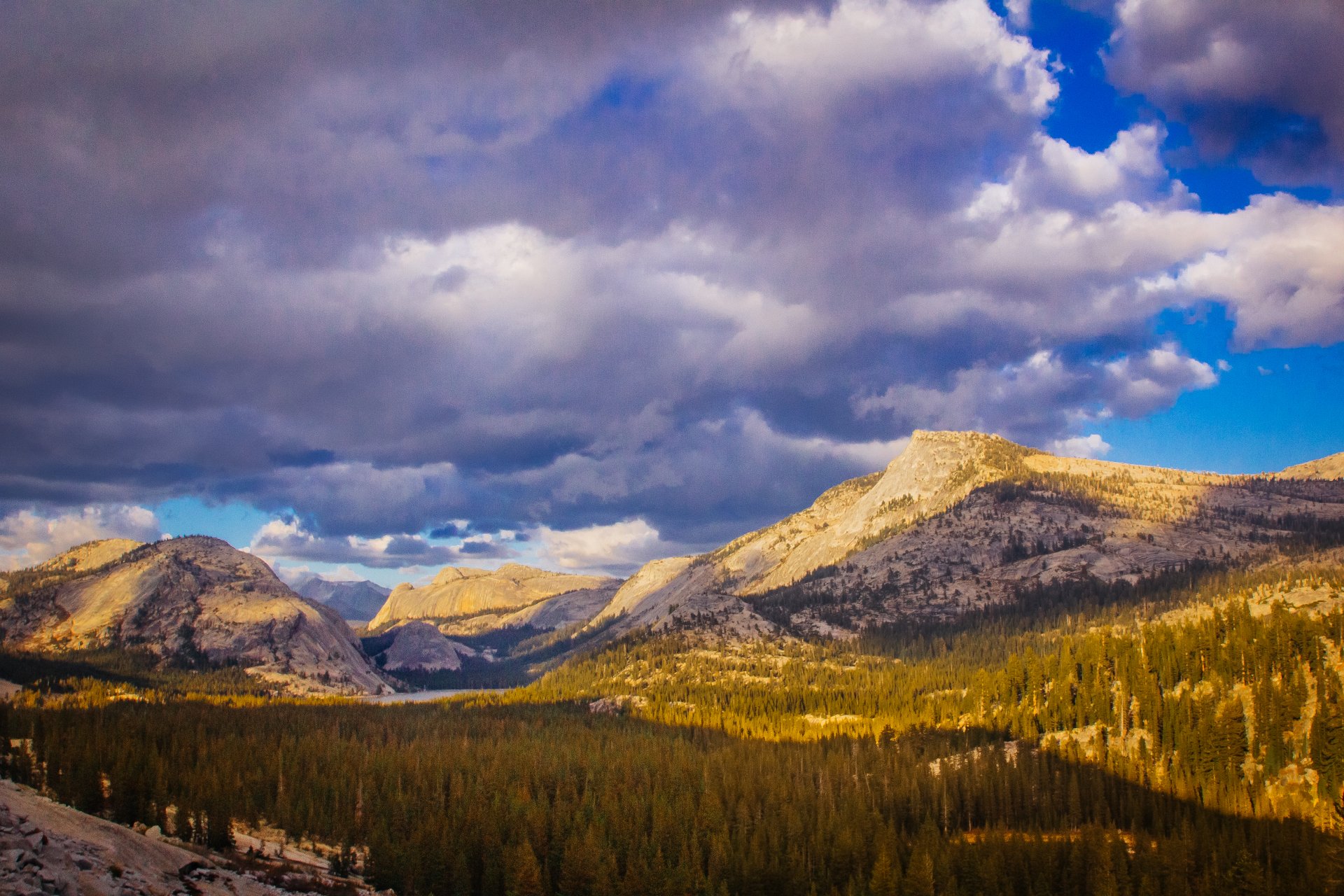 4K Ultra HD PC desktop wallpaper of a mountain landscape: sunlit peaks above a dense forest under dramatic cloud-filled sky, a vivid nature scene.