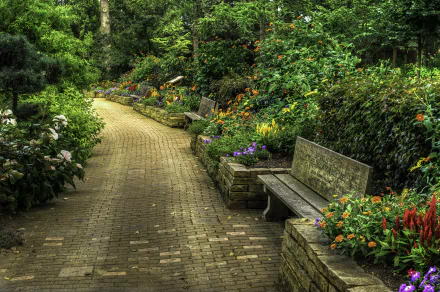 A serene stone walkway in a vibrant park, lined with blooming flowers, invites a tranquil spring stroll. Lush greenery and benches create a peaceful atmosphere. High-definition photography.