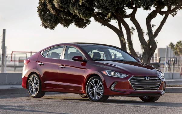 HD desktop wallpaper of a red Hyundai Elantra compact car parked outdoors near trees, showcasing the vehicle’s sleek design and modern features.