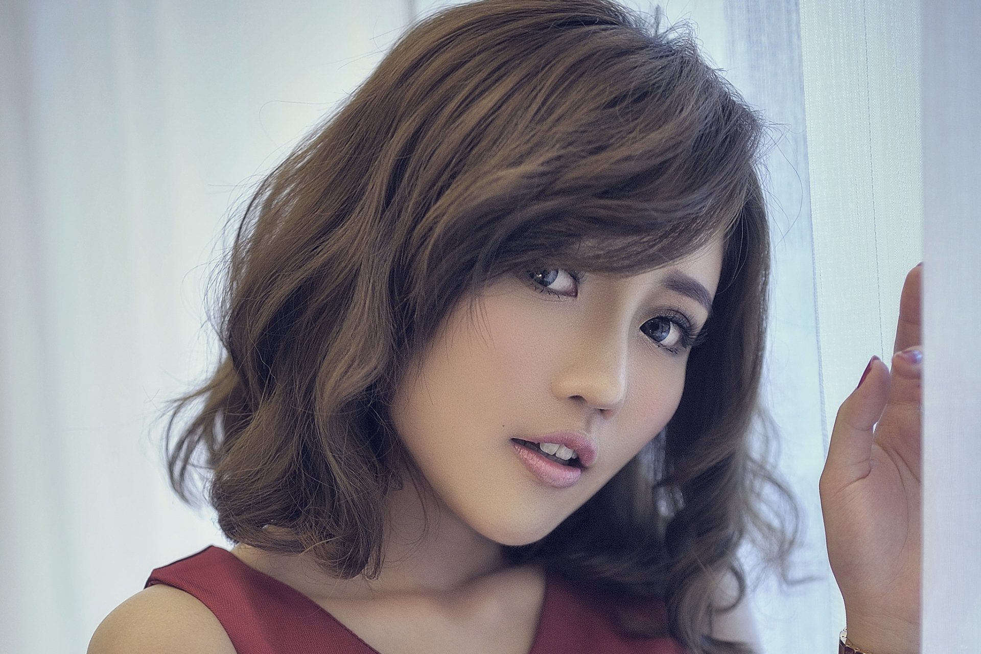 A stunning brunette Asian model with wavy hair and expressive eyes poses by a light curtain. This HD desktop wallpaper and background captures her in an elegant and natural moment.