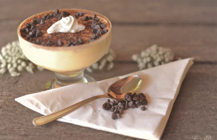 HD desktop wallpaper featuring a creamy tiramisu dessert topped with chocolate chips and whipped cream, accompanied by a spoon and scattered chocolate chips on a napkin.