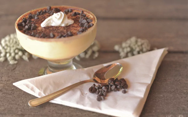 HD desktop wallpaper featuring a creamy tiramisu dessert topped with chocolate chips and whipped cream, accompanied by a spoon and scattered chocolate chips on a napkin.