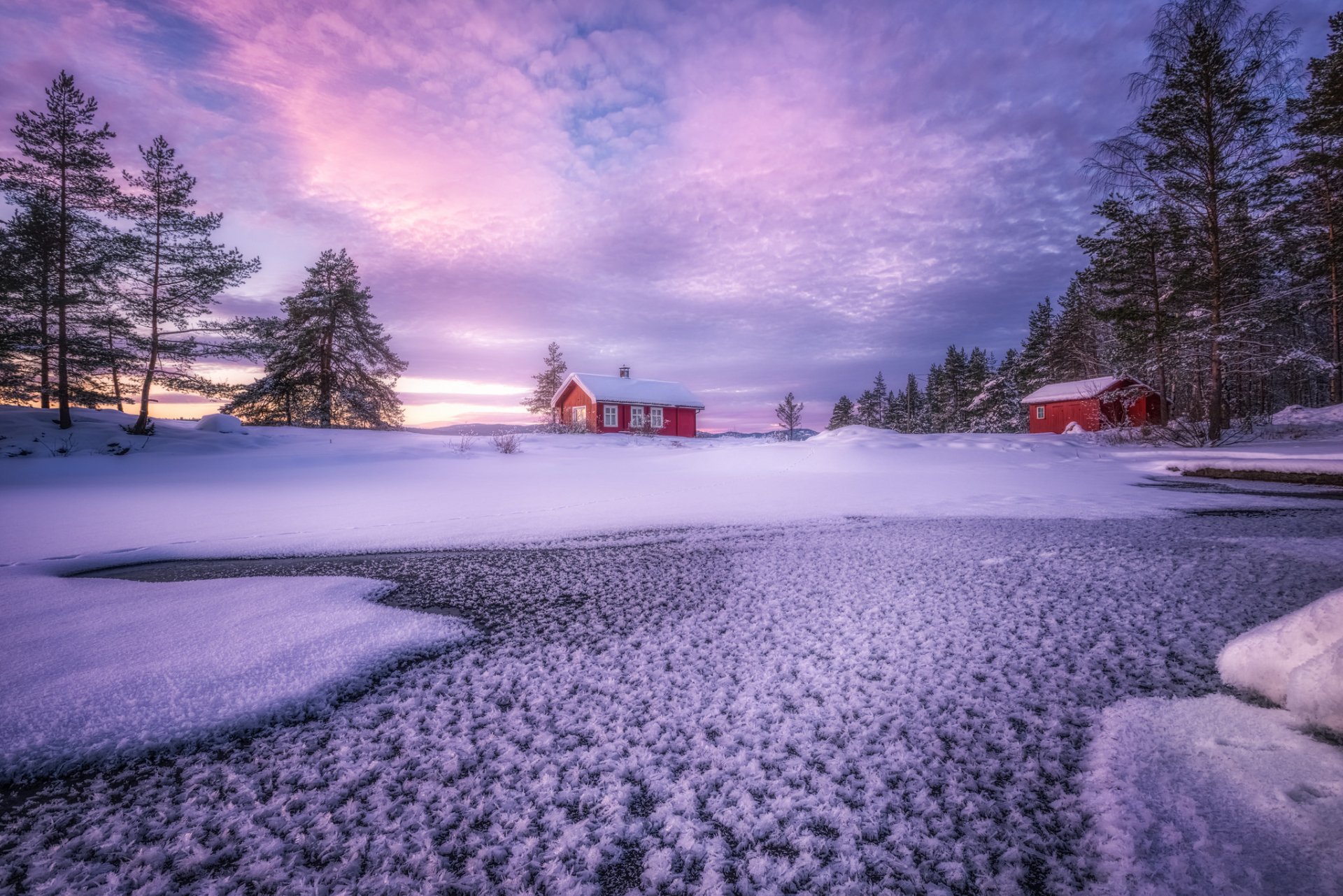 A serene winter HD desktop wallpaper featuring a snow-covered lake, tall trees, and man-made red houses under a colorful twilight sky.