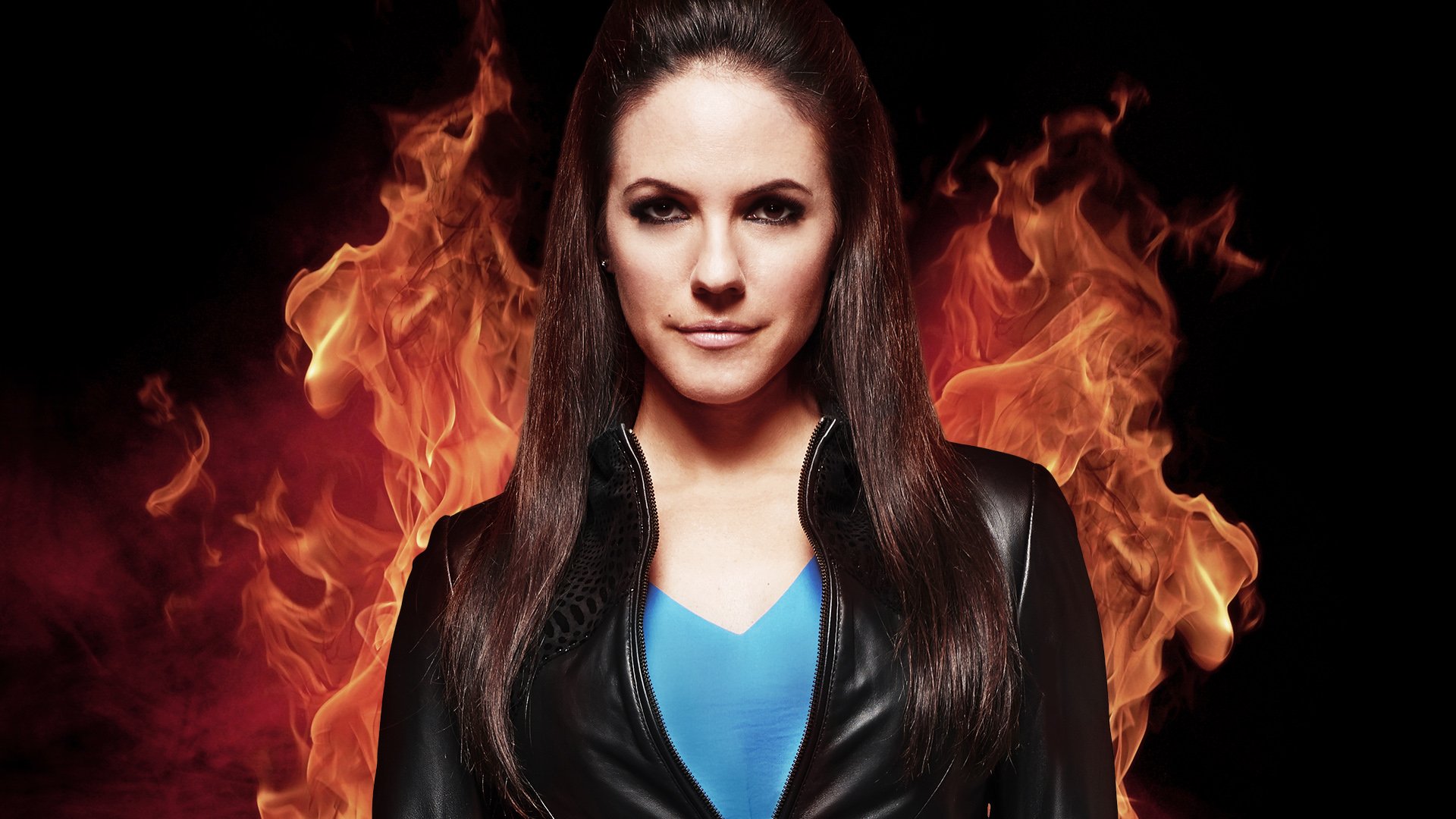 HD PC desktop wallpaper and background of a female character from the TV show Lost Girl against roaring flames, wearing a black jacket and blue top, staring confidently.