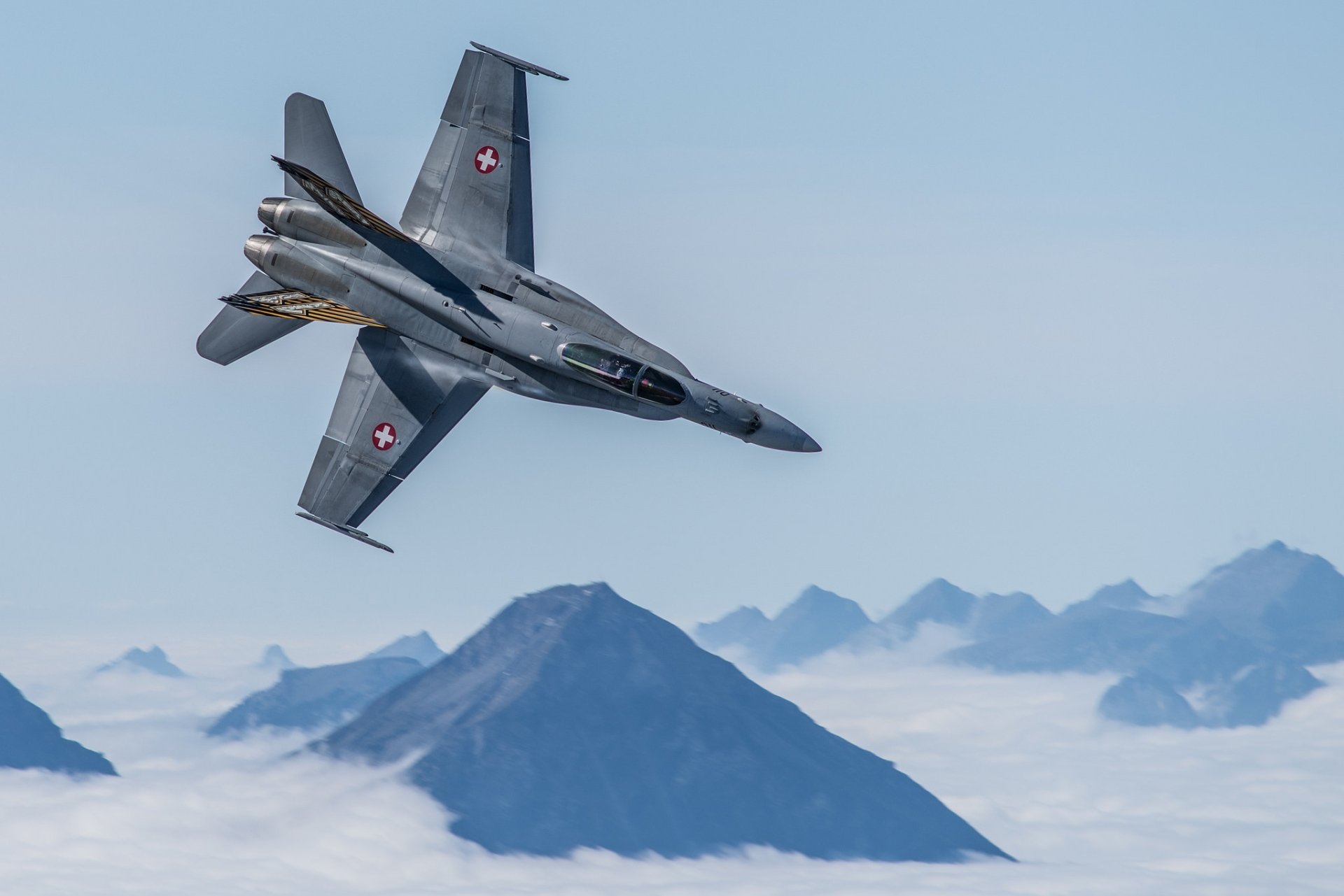 HD desktop wallpaper featuring a McDonnell Douglas F/A-18 Hornet jet fighter warplane soaring above clouds with mountain peaks in the background.