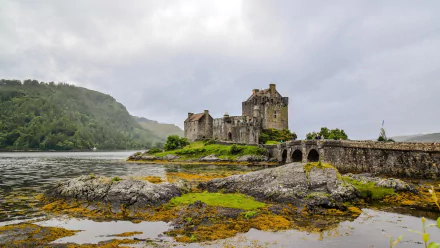 Scenic view of Eilean Donan Castle in Scotland, surrounded by a tranquil lake and rocky landscape, with a charming bridge leading to the castle, under a cloudy sky.