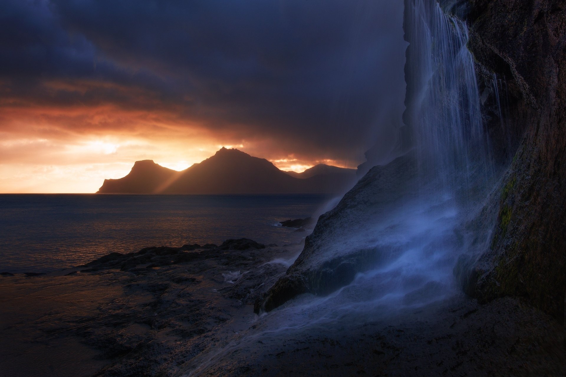 HD desktop wallpaper of a serene coastal waterfall cascading into the rocky shore at sunset, with silhouetted mountains in the background and a vibrant sky.