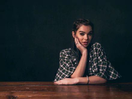 A brunette American actress, Hailee Steinfeld, poses thoughtfully with her chin resting on her hand, showcasing a stylish plaid shirt against a dark background.