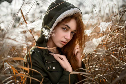 A redhead woman with hazel eyes wearing a hooded jacket, surrounded by dry plants, captured in a striking HD desktop wallpaper.