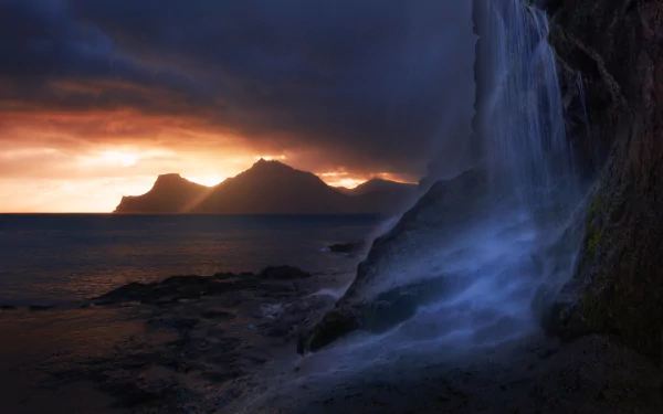 A serene coastal scene featuring a stunning waterfall cascading over rocks, framed by mountains under a dramatic sunset sky, perfect for a nature-inspired HD desktop wallpaper.