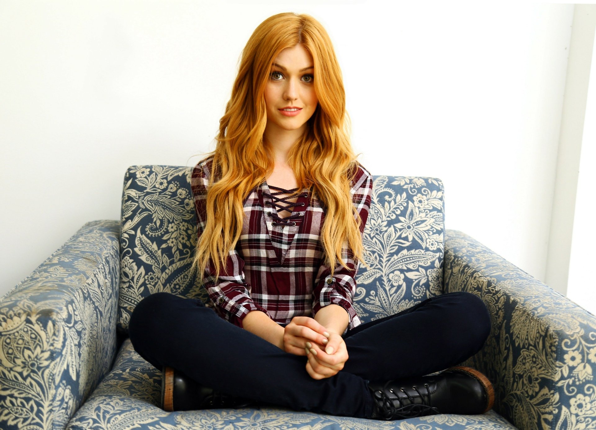 Katherine McNamara, an American actress with striking green eyes and red hair, poses casually on a patterned couch, showcasing her vibrant style in this HD desktop wallpaper.