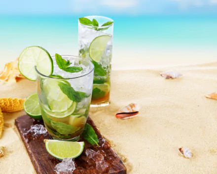 Refreshing mojito cocktails with ice, lime, and mint on sandy beach with seashells, captured in vibrant 4K Ultra HD summer desktop wallpaper and background.