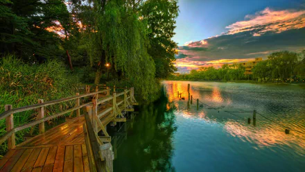 A serene boardwalk surrounded by lush green trees stretches along a calm lake under a colorful sunset sky, captured in vibrant HD photography.